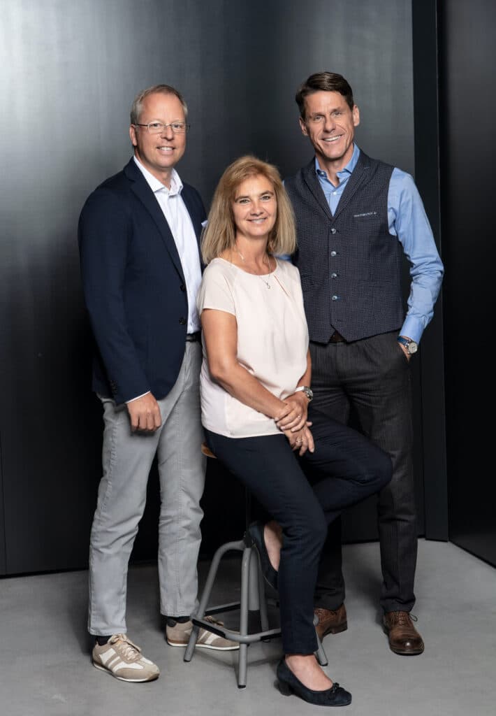 Drei Personen stehen und sitzen in einem modernen Studio, lächelnd in die Kamera blickend. Zwei Männer tragen Blazer, Hemden und Freizeitschuhe, während die Frau auf einem hohen Hocker sitzt, gekleidet in ein helles Oberteil und dunkle Hosen. Der Hintergrund ist dunkel und schlicht. Von links nach rechts, Herr Markus Glöggler, Frau Stephanie Sattler-Rick, Herr Jürgen Kohler