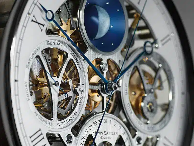 Detailaufnahme des komplizierten Uhrwerks der Opus Perpetual von Erwin Sattler mit sichtbaren Zahnrädern, blauen Zeigern und Mondphasenanzeige. Die Tage der Woche sind auf kreisförmigen Anzeigen sichtbar. Das Uhrwerk zeichnet sich durch seinen ewigen Kalender aus