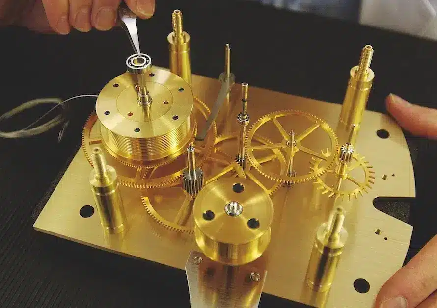 Mechanismus aus goldfarbenem Metall mit mehreren Zahnrädern; Hände justieren ein Bauteil mit einer Pinzette.