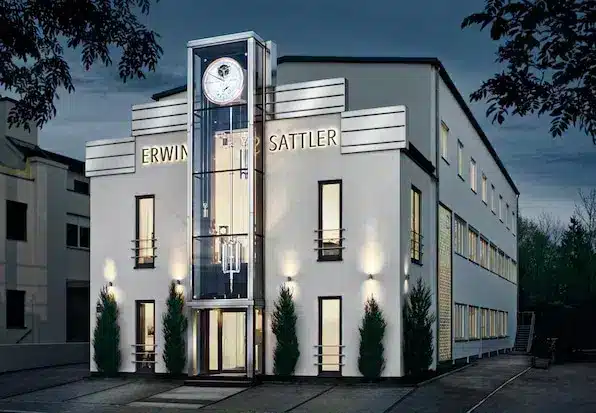 Modernes Gebäude mit gläsernem Eingangsturm und Uhr, beschriftet mit "Erwin Sattler". Beleuchtete Fassade und umgeben von Bäumen im Abendlicht.