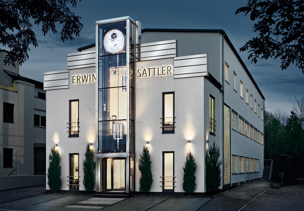 Modernes Gebäude mit gläsernem Eingangsturm und Uhr, beschriftet mit "Erwin Sattler". Beleuchtete Fassade und umgeben von Bäumen im Abendlicht.