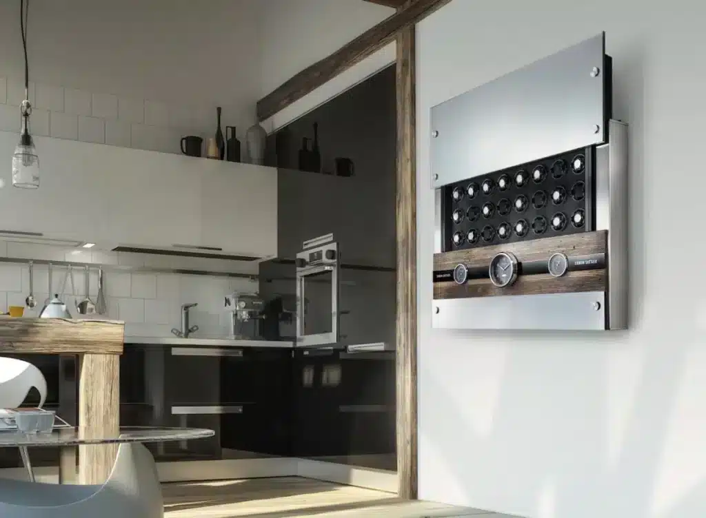 Uhrenbeweger Rotalis 24 von Erwin Sattler hängt an einer hellen Wand. Oberes Glaspanel ist offen und die 24 Uhrenbeweger sind sichtbar. In der Mitte befindet sich ein Altholz Panel mit zwei nautischen Instrumenten (Thermometer und Barometer) sowie eine Quarzuhr. alle haben ein schwarzes Zifferblatt. Hinter einer geöffneten Klappe sieht man noch den Stauraum für Ringe oder Armbänder. Im Hintergrund sieht man eine moderne Küche mit weißer und schwarzer Einrichtung.