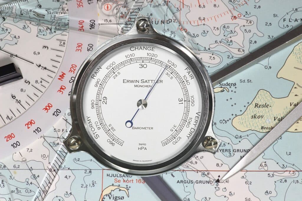 Analoger Barometer von Erwin Sattler auf einer Seekarte, zeigt Luftdruck in hPa und inHg an, umgeben von Navigationswerkzeugen.