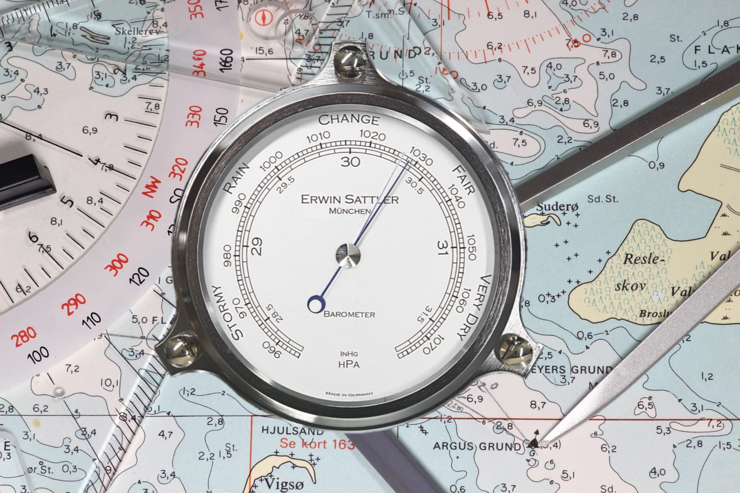 Analoger Barometer von Erwin Sattler auf einer Seekarte, zeigt Luftdruck in hPa und inHg an, umgeben von Navigationswerkzeugen.