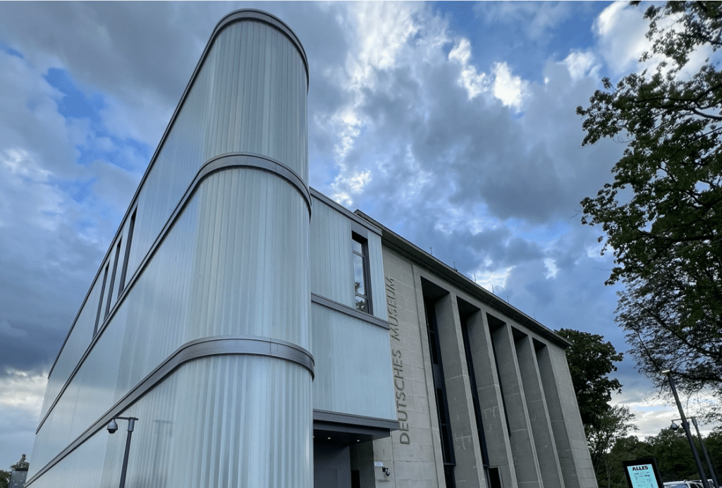 Ein modernes Gebäude mit gerundeten Kanten und vertikalen Fensterreihen, an dem der Schriftzug „Deutsches Museum“ angebracht ist. Der Himmel ist bewölkt, Bäume sind auf der rechten Seite sichtbar.