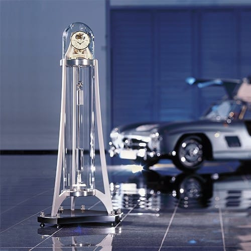 Eine moderne Standuhr mit Glasgehäuse, die Columna Temporis von Erwin Sattler im Vordergrund, im Hintergrund ein klassisches Auto mit offenem Flügeltürendesign in einer hellen Halle.