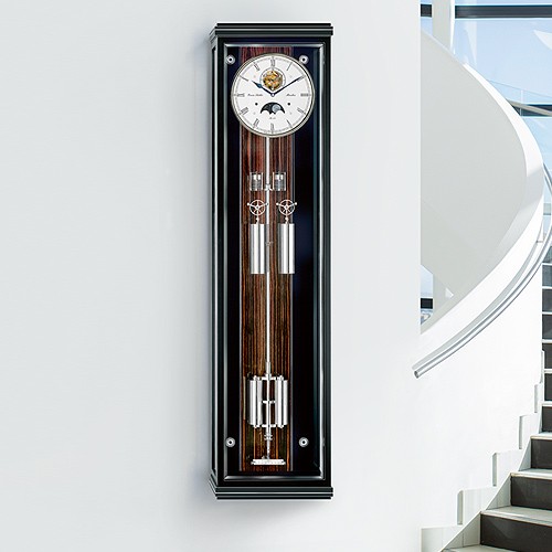 Elegante Pendeluhr Secunda Sonata von Erwin Sattler mit großem weißen Zifferblatt und mehreren glänzenden Metallkomponenten, montiert an einer Innenwand neben einer modernen Treppe.