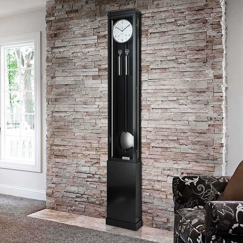 Schwarze Standuhr Excelsia von Erwin Sattler vor einer rustikalen Steinwand, daneben ein Sessel mit gemustertem Bezug, in einem hell beleuchteten Raum.