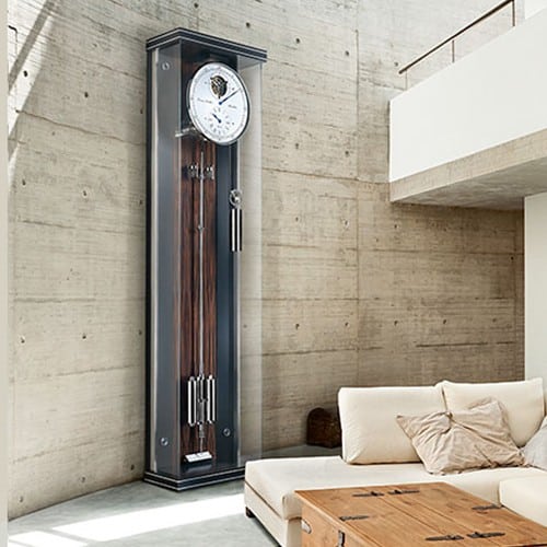 Standuhr Maxima Secunda von Erwin Sattler in modernem Wohnzimmer mit Betonwand, neben einem cremefarbenen Sofa und Holztisch.