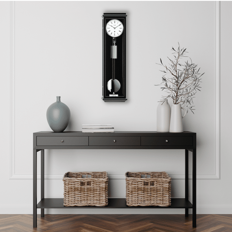 Schwarze Kommode mit drei Schubladen, darauf eine graue Vase, ein Stapel Bücher und zwei weiße Vasen mit Zweigen. An der Wand hängt eine schwarze Classica 60 von Erwin Sattler. Darunter stehen zwei geflochtene Körbe. Holzboden mit Fischgrätmuster.