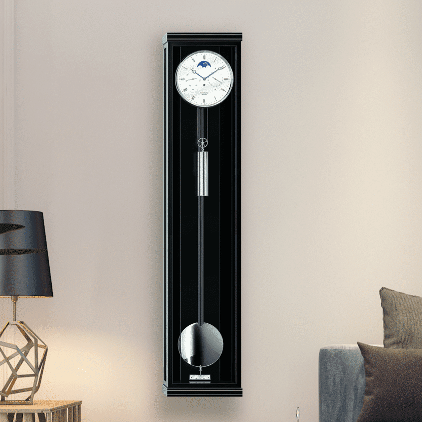 Eine elegante, schwarze Wanduhr Classica K130M mit Pendel und Mondphasenanzeige vor einer beigen Wand. Links ist teilweise eine moderne Tischlampe und rechts ein graues Sofa erkennbar.