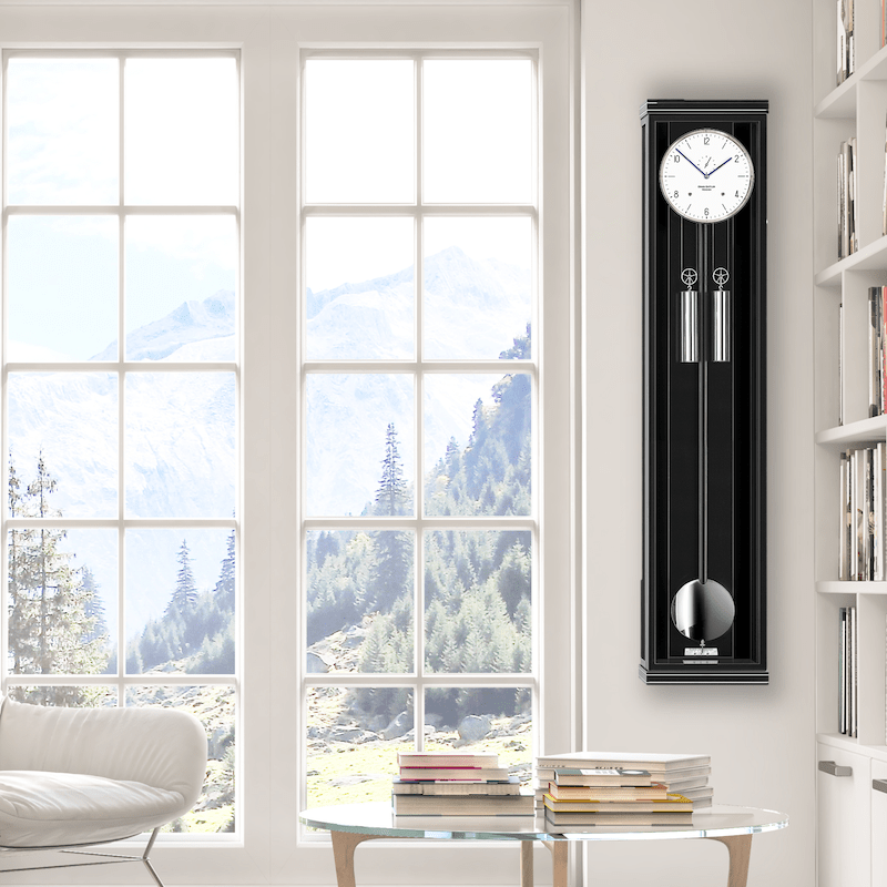 Helles Wohnzimmer mit einer großen Fensterfront, die eine Berglandschaft zeigt; an der Wand eine schwarze Pendeluhr; Modell Classica S130M von Erwin Sattler in einer modernen Einrichtung mit Büchern auf einem runden Glastisch.