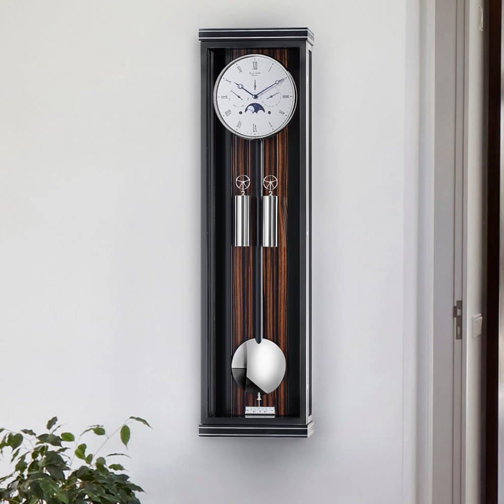 Wanduhr Classica KS100M von Erwin Sattler mit Pendel in schwarz und Makassar Rückwand in einem schmalen, modernen Gehäuse an einer weißen Wand. Unten sind Pflanzen zu sehen.