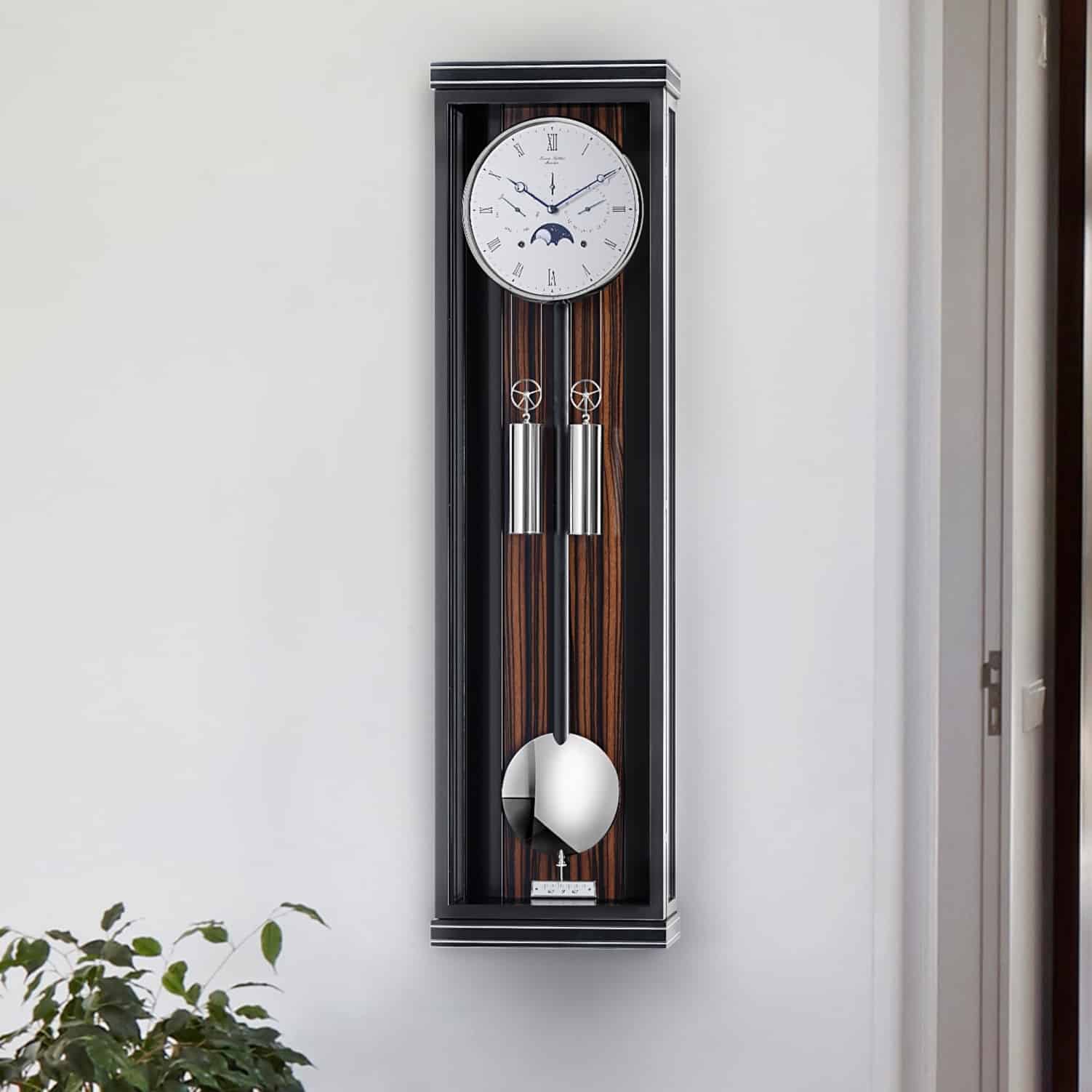 Wanduhr Classica KS100M von Erwin Sattler mit Pendel in schwarz und Makassar Rückwand in einem schmalen, modernen Gehäuse an einer weißen Wand. Unten sind Pflanzen zu sehen.