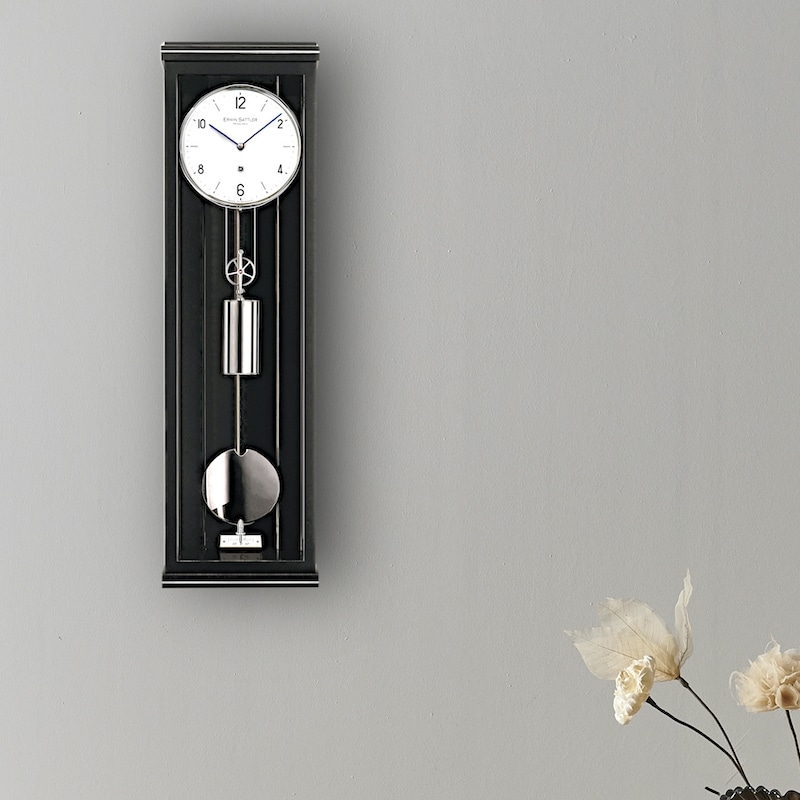 Wanduhr mit Pendel in schwarzem Gehäuse, darunter eine Vase mit getrockneten Blumen, vor einer grauen Wand, Modell Classica P70M von Erwin Sattler.