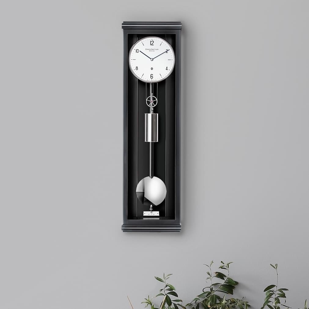 Wanduhr mit Pendel in schwarzem Gehäuse, darunter eine Vase mit Blumen, vor einer grauen Wand, Modell Classica P70M von Erwin Sattler.