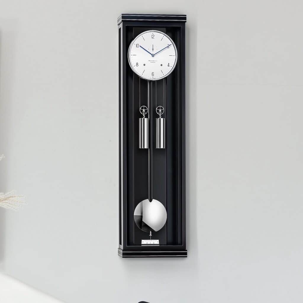 Wanduhr Classica S00 von Erwin Sattler in einem schwarzen Gehäuse mit zwei silbernen Gewichten und einem Pendel, montiert an einer hellgrauen Wand.