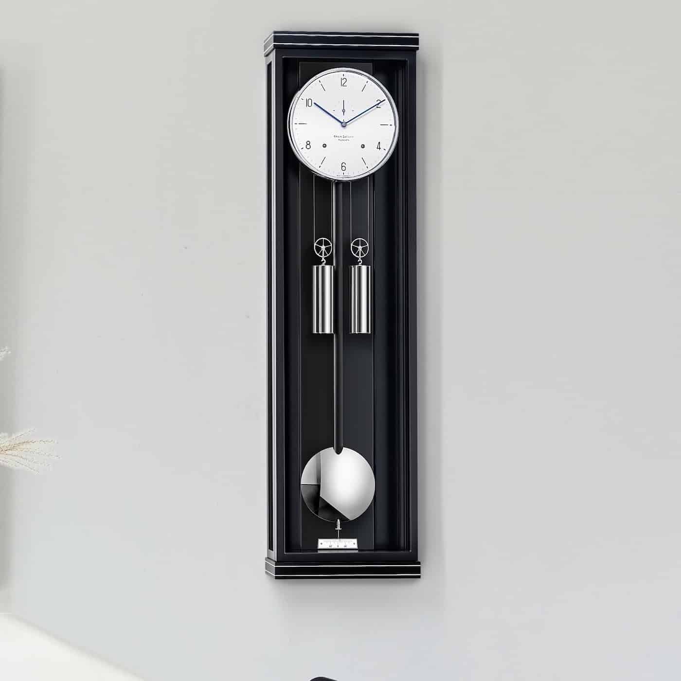 Wanduhr Classica S00 von Erwin Sattler in einem schwarzen Gehäuse mit zwei silbernen Gewichten und einem Pendel, montiert an einer hellgrauen Wand.