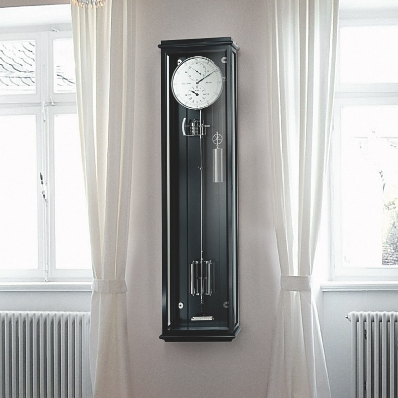 Wanduhr Classica Secunda 1985 von Erwin Sattler im klassischen Design mit schwarzem Gehäuse und sichtbarem Pendel, zwischen zwei großen Fenstern mit weißen Vorhängen.