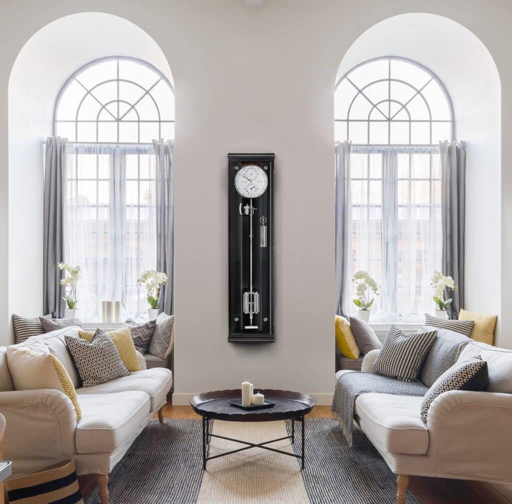 Wanduhr Classica Secunda 1985 von Erwin Sattler im klassischen Design mit schwarzem Gehäuse und sichtbarem Pendel, zwischen zwei großen Fenstern mit weißen Vorhängen.