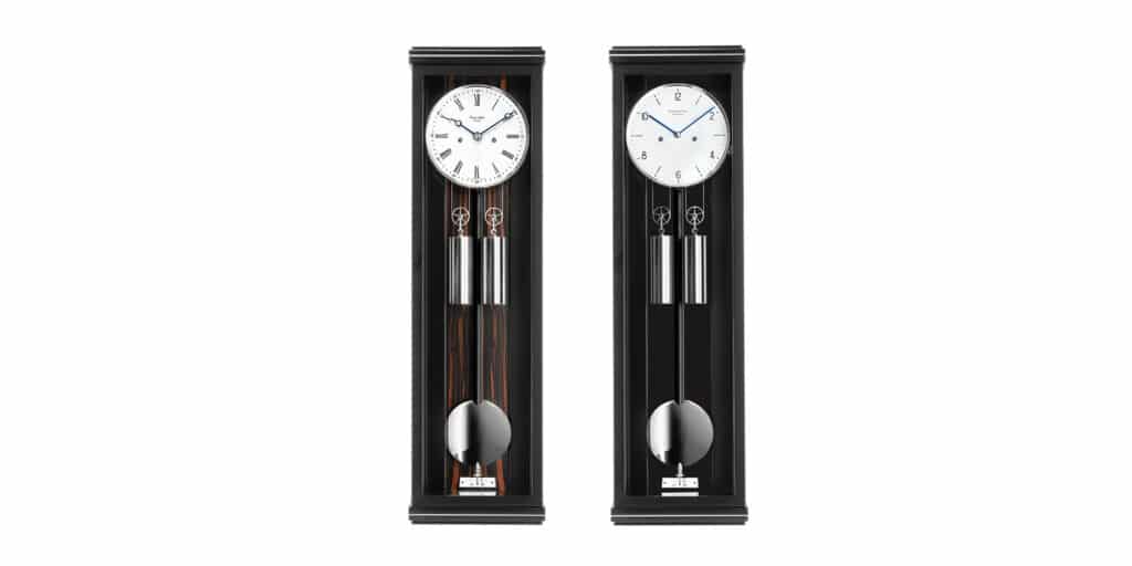 Zwei Wandpendeluhren, Classica S70M von Erwin Sattler mit hellem Zifferblatt einmal mit arabischen und einmal mit römischen Ziffern auf dem Zifferblatt, blauen Zeigern und Mondphasenanzeige. Jede Uhr hat zwei Gewichte und ein Pendel. Links in schwarz mit Makassar Rückwand, rechts in ganz schwarz.