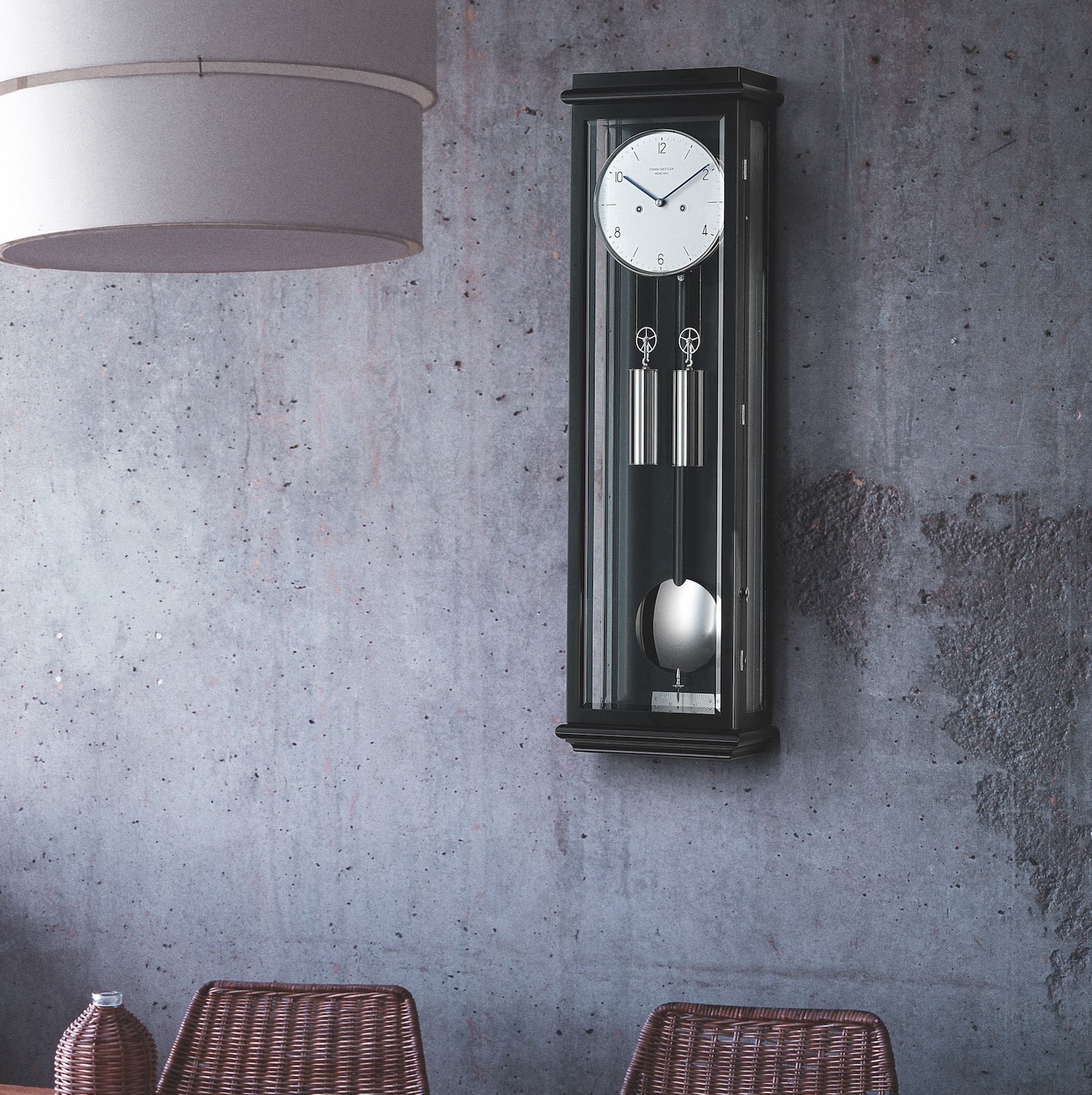 Wanduhr mit Pendel in schwarz, Modell Classica S70 von Erwin Sattler an einer grauen Betonwand, daneben ein Tisch mit geflochtenen Stühlen.