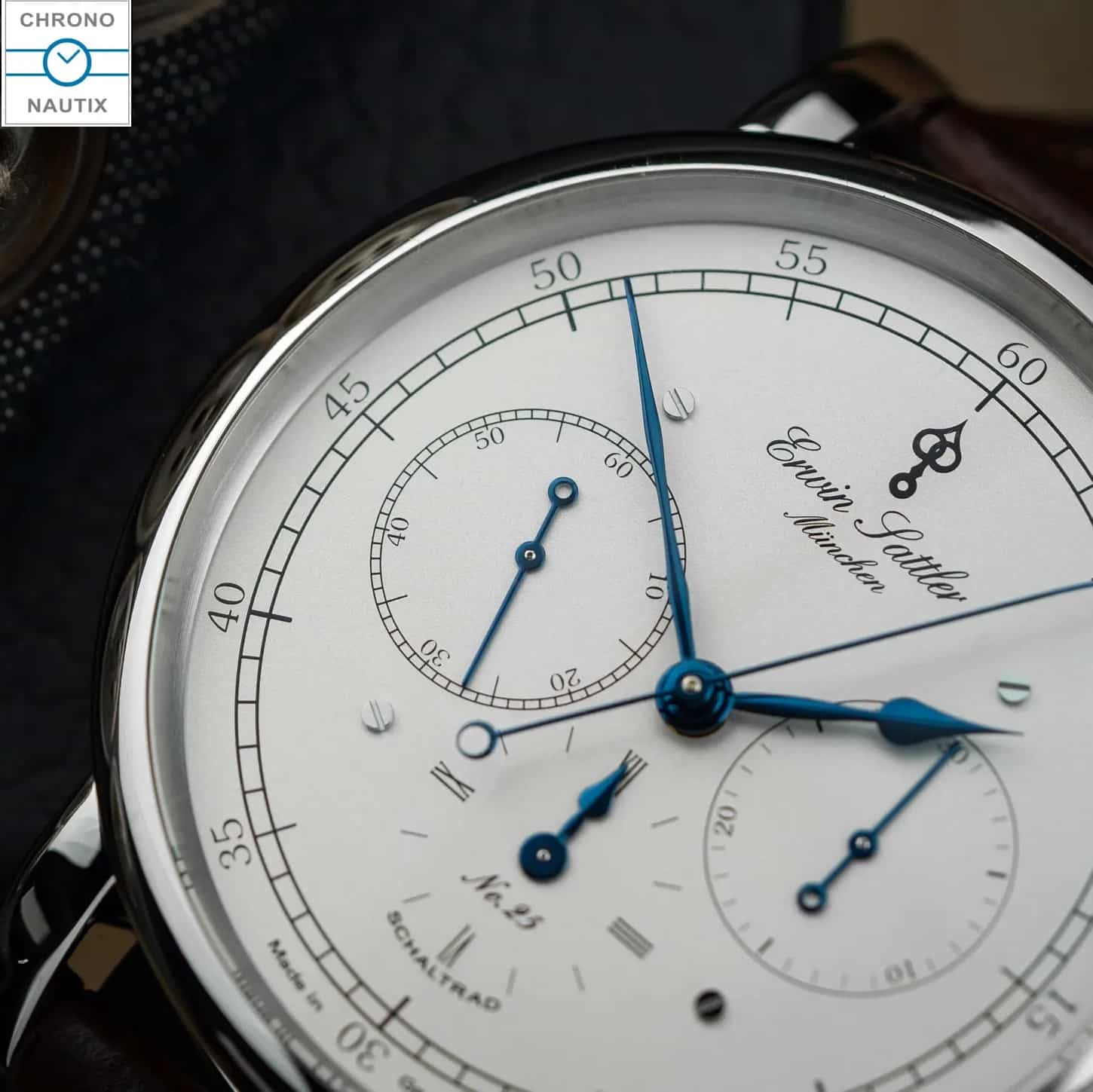 Nahaufnahme einer eleganten Armbanduhr mit weißem Zifferblatt, blauen Zeigern und detaillierter Skala, die die Inschrift „Erwin Sattler, München“ trägt.