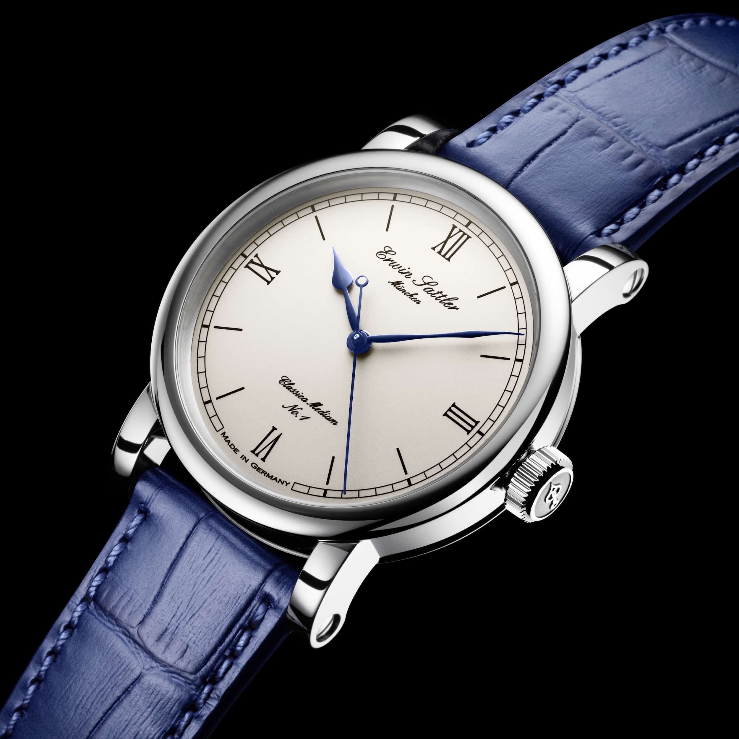 Armbanduhr Classica Medium von Erwin Sattler mit silbernem Gehäuse, weißem Zifferblatt, blauen Zeigern und römischen Ziffern, getragen an einem blauen Lederarmband.