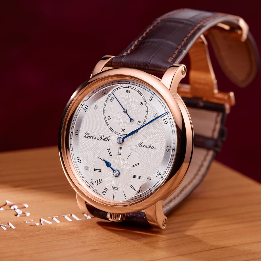 Armbanduhr Regulateur 1920 von Erwin Sattler mit roségoldenem Gehäuse und braunem Lederarmband, auf einem Holztisch liegend. Zifferblatt mit zwei Hilfszifferblättern und blauen Zeigern. Text auf Zifferblatt: "Erwin Sattler München".