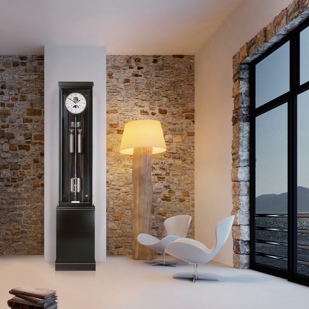 Standuhr Troja Sonata  von Erwin Sattler mit Glasfront in modernem Wohnzimmer, daneben eine rustikale Stehlampe mit Holzfuß und zwei weiße Designerstühle vor einer Steinwand.