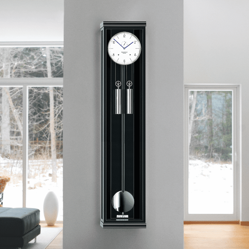 Helles Wohnzimmer mit einer großen Fensterfront, die eine Berglandschaft zeigt; an der Wand eine schwarze Pendeluhr; Modell Classica S130M von Erwin Sattler in einer modernen Einrichtung mit Büchern auf einem runden Glastisch.