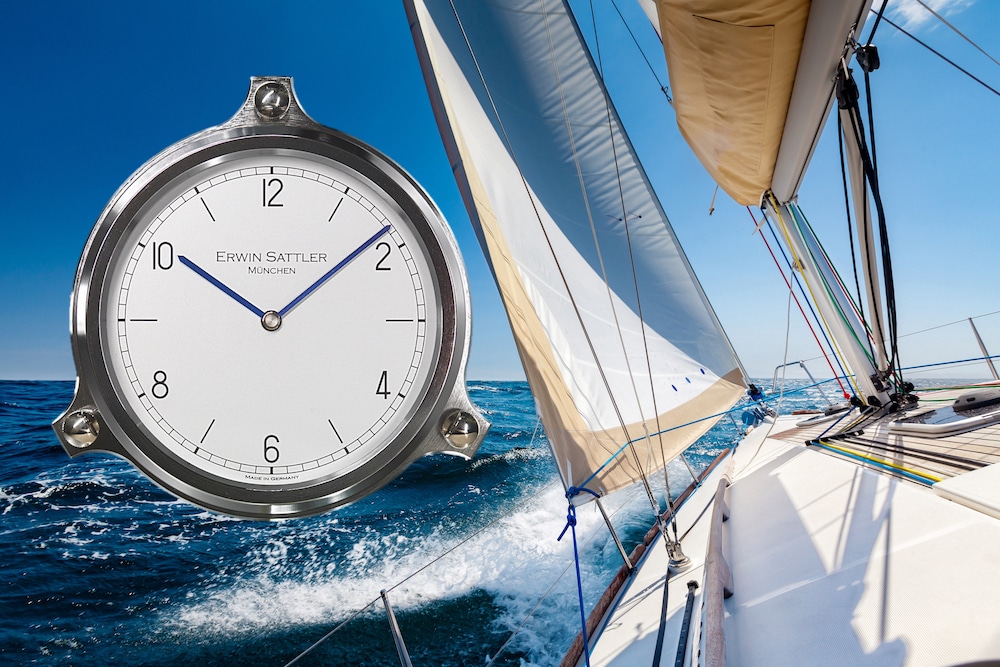 Eine runde Uhr, Modell Navalis von Erwin Sattler, mit blauem Stunden- und Minutenzeiger sowie einem weißen Zifferblatt ist überlagert auf einem Bild, das ein Segelboot auf offener See zeigt. Das Segelboot segelt bei klarem Himmel und die Sonne wirft Schatten auf das Deck.