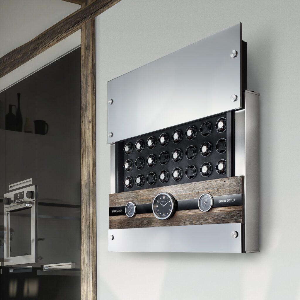 Uhrenbeweger Rotalis 24 von Erwin Sattler hängt an einer hellen Wand. Oberes Glaspanel ist offen und die 24 Uhrenbeweger sind sichtbar. In der Mitte befindet sich ein Altholz Panel mit zwei nautischen Instrumenten (Thermometer und Barometer) sowie eine Quarzuhr. alle haben ein schwarzes Zifferblatt. Hinter einer geöffneten Klappe sieht man noch den Stauraum für Ringe oder Armbänder. Im Hintergrund sieht man eine moderne Küche mit weißer und schwarzer Einrichtung.