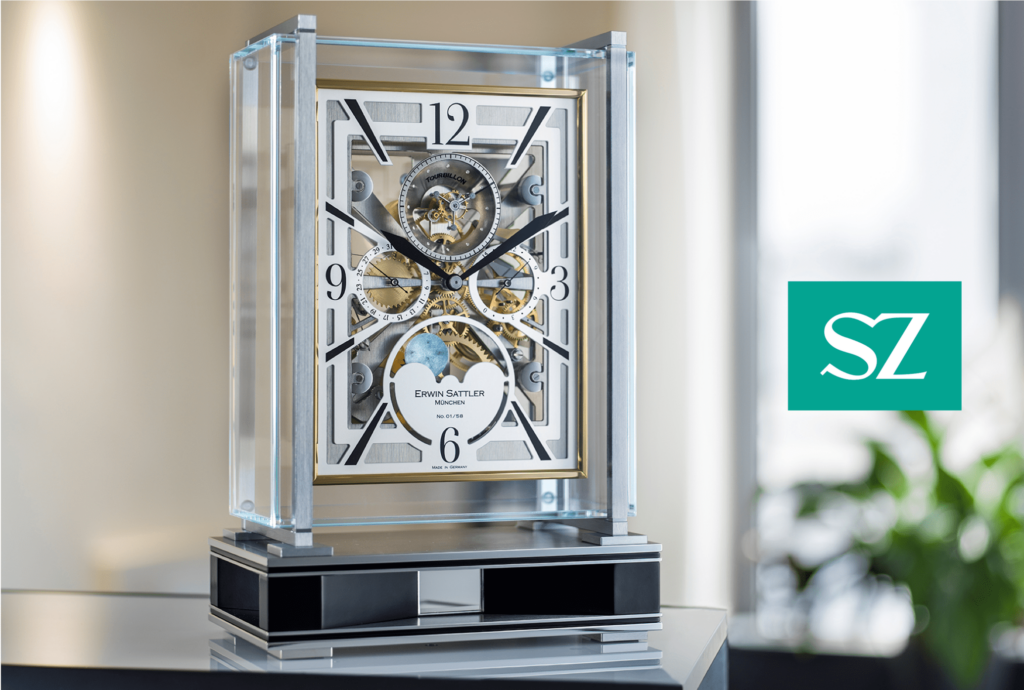 Eine moderne, durchsichtige Tischuhr, Opus Tourbillon von Erwin Sattler mit sichtbarem Uhrwerk und goldenen Details steht auf einem Spiegel; sie zeigt die Uhrzeit 10:10 an. Ein Logo mit den Buchstaben "SZ" ist auf der rechten Seite zu sehen. Im Hintergrund befindet sich eine unscharfe Pflanze.