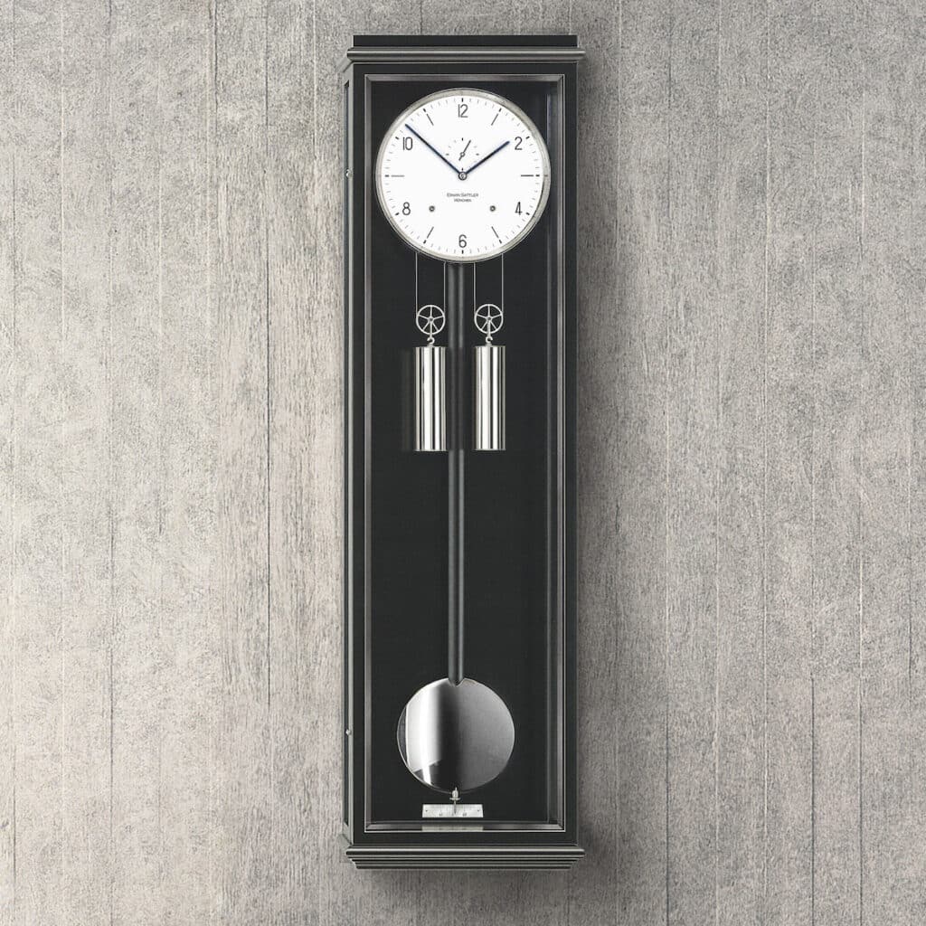 Eine moderne Wanduhr mit Pendel und Gewichten in einem schlanken schwarzen Gehäuse vor einer grauen Holzwand, Modell Classica S100 von Erwin Sattler.