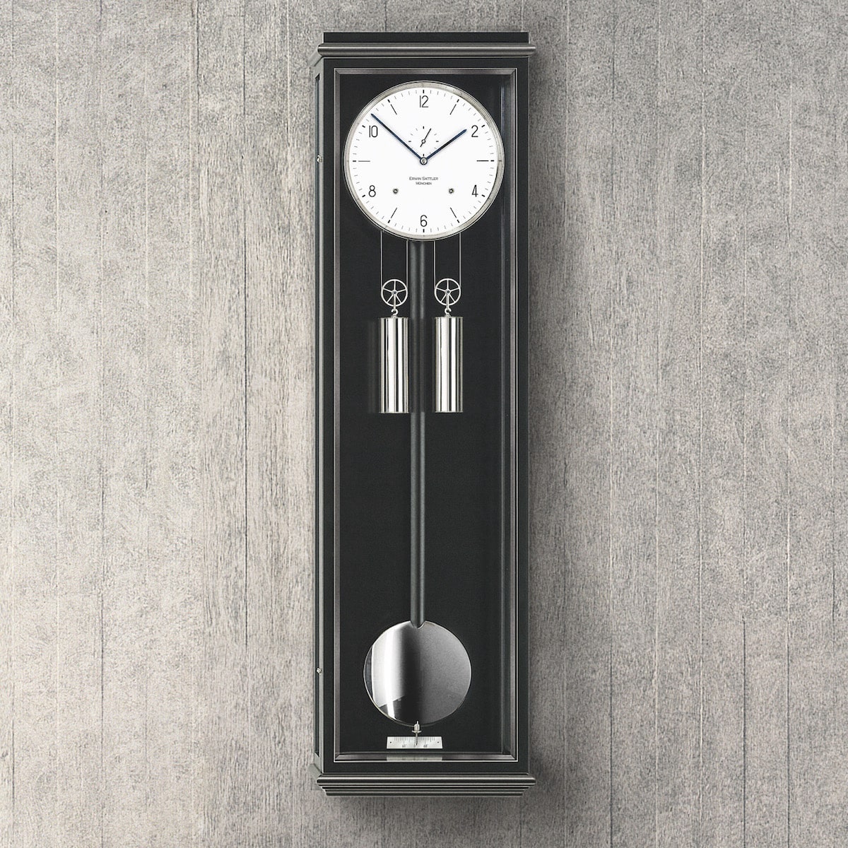 Eine moderne Wanduhr mit Pendel und Gewichten in einem schlanken schwarzen Gehäuse vor einer grauen Holzwand, Modell Classica S100 von Erwin Sattler.