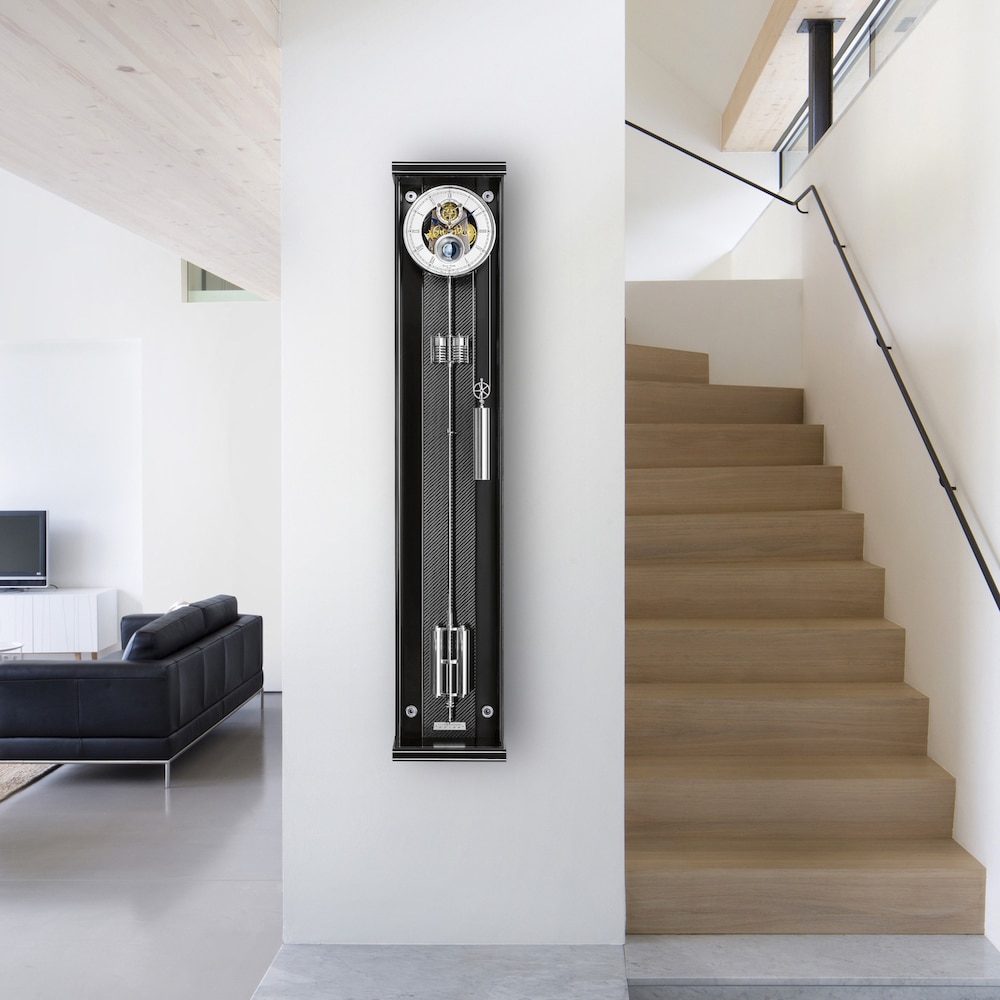 Moderne Wohnzimmeransicht mit einer hölzernen Treppe auf der rechten Seite und einer schwarzen Wandpendeluhr , Modell Opus Secunda 130 M von Erwin Sattler, an einer weißen Wand auf der linken Seite. Im Hintergrund befindet sich eine schwarze Ledercouch und ein Fernseher.