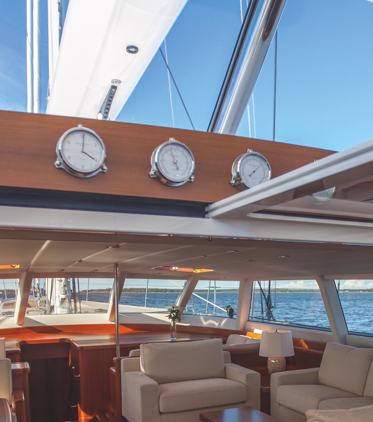 Innenansicht einer Yacht mit modernen, beigen Sofas und Holzmöbeln, großen Fenstern mit Blick auf das Meer und drei nautischen Instrumenten von Erwin Sattler auf einem Holzpanel befestigt.