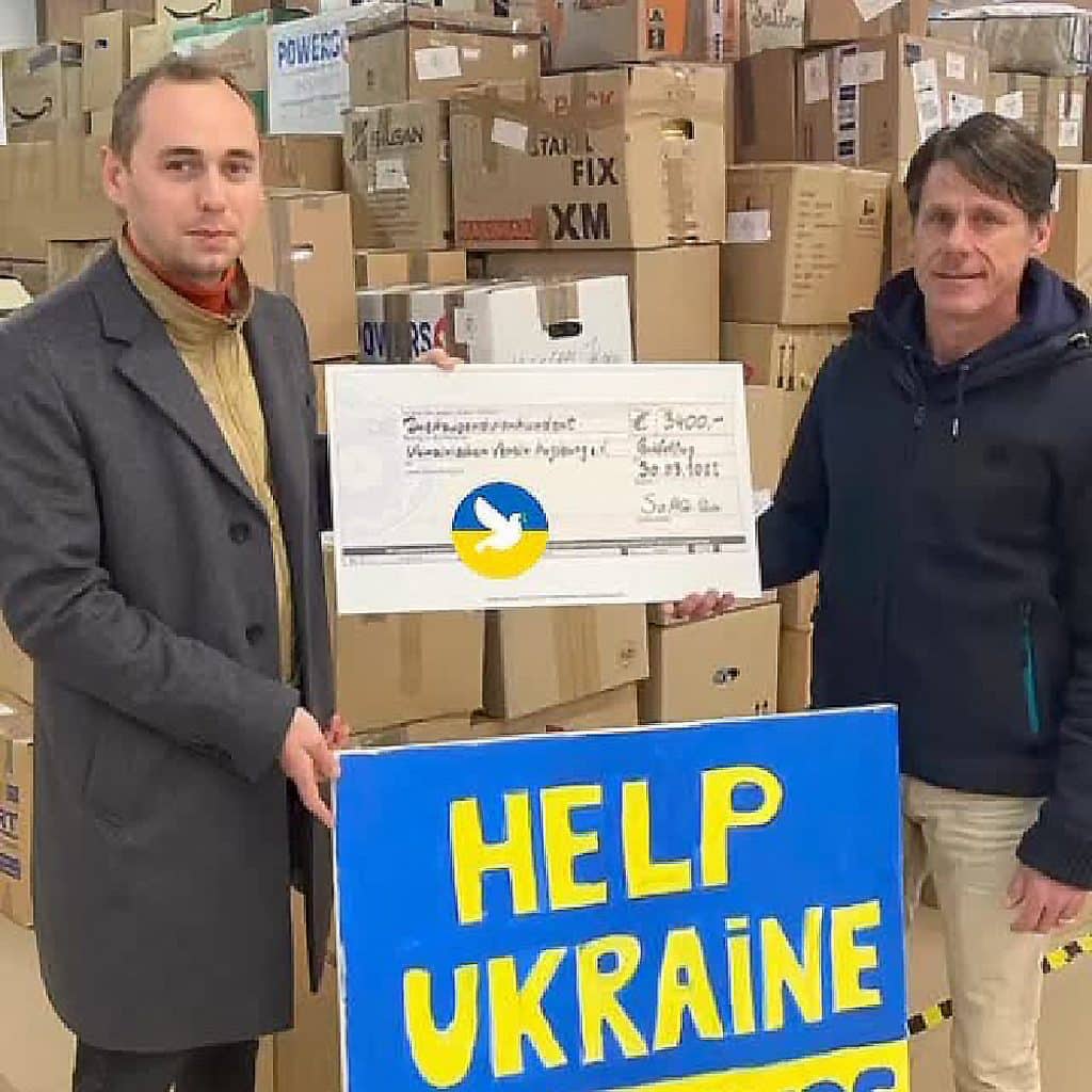 Zwei Männer halten einen symbolischen Scheck und ein blaues Schild mit gelber Aufschrift „HELP UKRAINE“. Im Hintergrund sind gestapelte Kartons zu sehen.