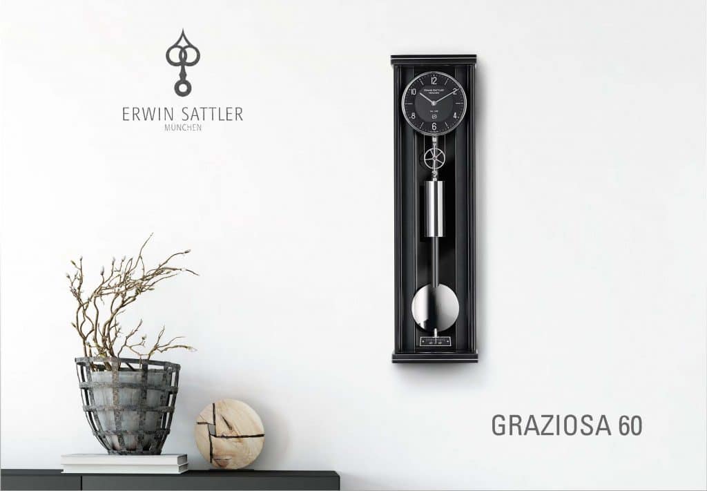 Wanduhr Graziosa 60 mit schwarzem, rechteckigem Gehäuse und silbernem Pendel, daneben ein Topf mit dürren Zweigen auf einem Tisch. Text "Erwin Sattler München" und "GRAZIOSA 60" an der Wand.