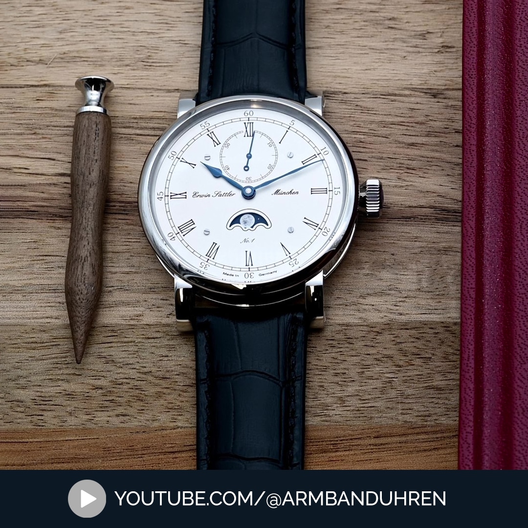 Armbanduhr Classica Lunaris von Erwin Sattler mit silbernem Zifferblatt, blauen Zeigern, römischen Ziffern und schwarzem Lederarmband. Enthält eine Mondphasenanzeige und eine separate kleine Sekundenanzeige, neben einem hölzernen Schreibgerät auf einem Holztisch.