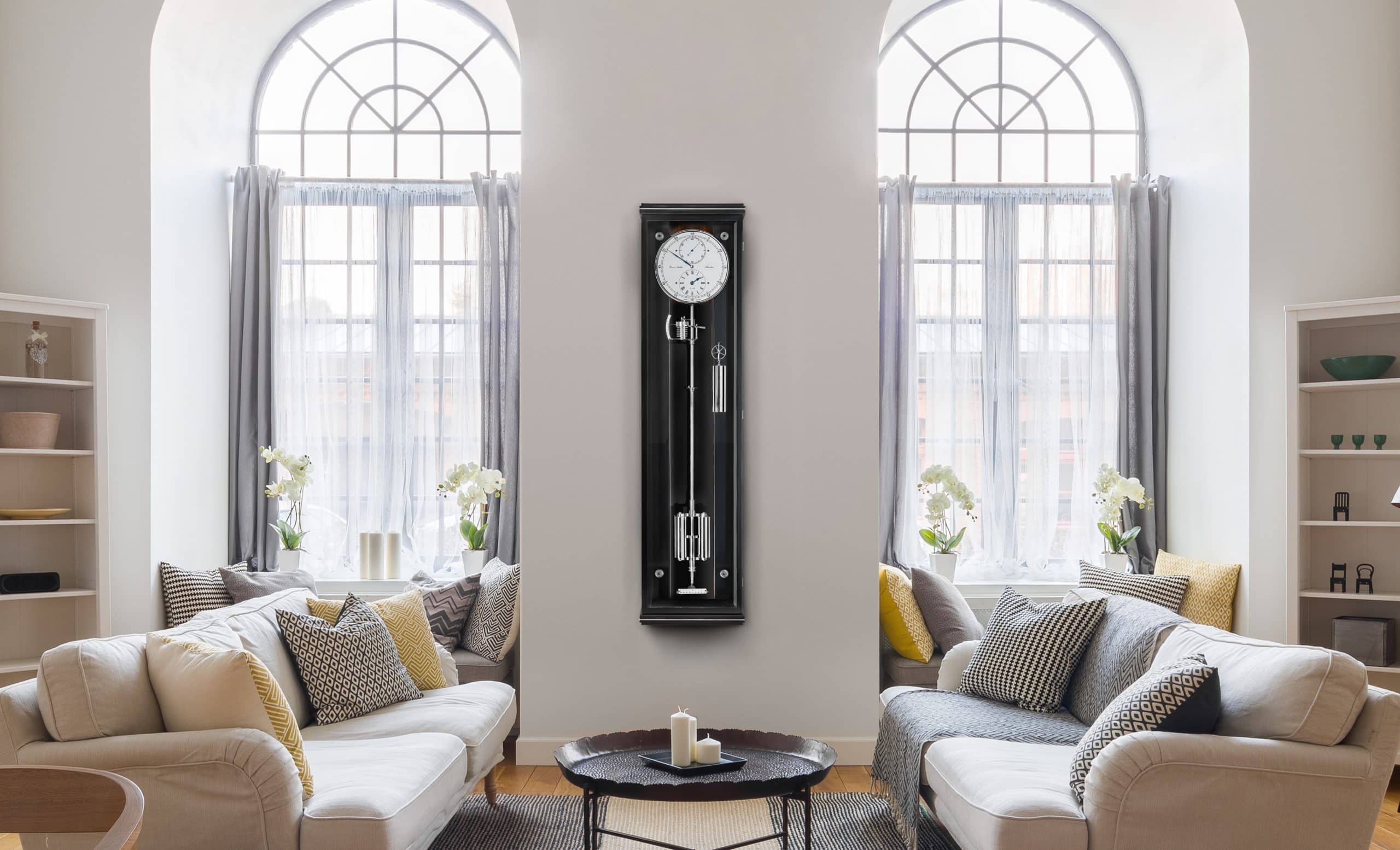 Wanduhr Classica Secunda 1985 von Erwin Sattler im klassischen Design mit schwarzem Gehäuse und sichtbarem Pendel, zwischen zwei großen Fenstern mit weißen Vorhängen.