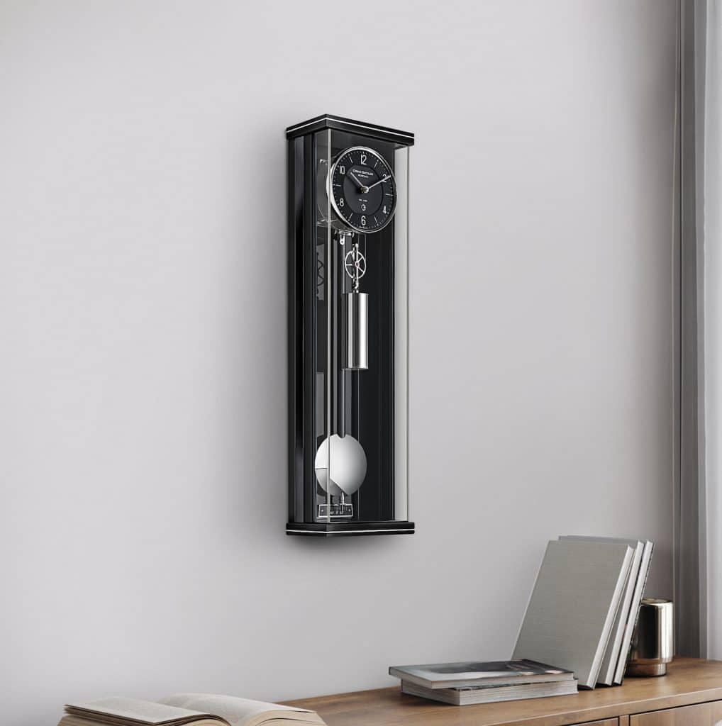 Schwarze Wanduhr mit einem Pendel, Modell Graziosa 60 von Erwin Sattler, das in einem modernen, minimalistisch eingerichteten Raum an der Wand hängt, daneben ein Tisch mit Büchern und einer Tasse.