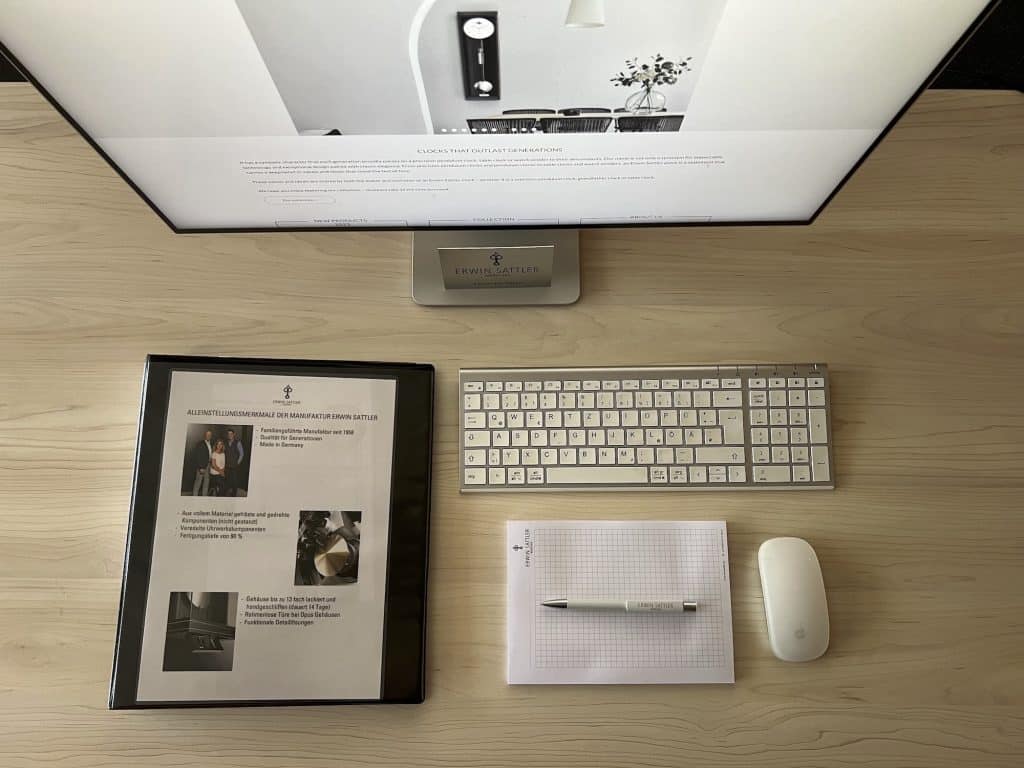 Arbeitsfläche mit Computerbildschirm, beleuchtetem Tastatur- und Mouse-Setup, Notizblock mit Stift sowie einer Broschüre auf einem hellen Holztisch. Bildschirm zeigt schwarz-weiße Innenaufnahme mit Pflanze.