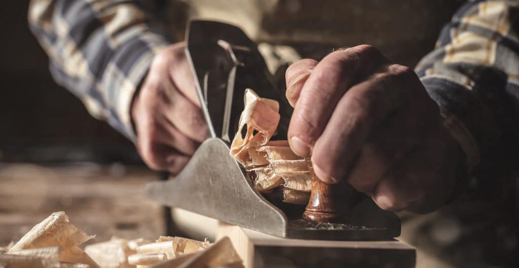 Eine Person hobelt ein Stück Holz, dabei fliegen Holzspäne in einer Werkstattumgebung.