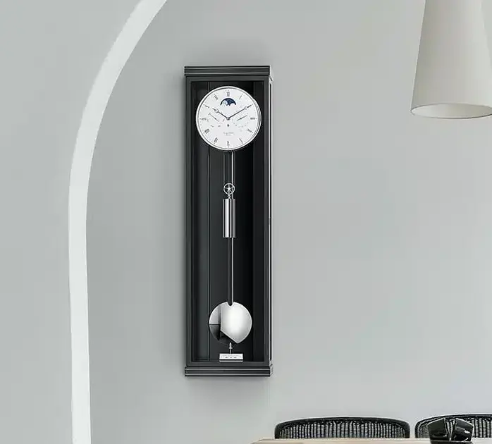 Eine Wanduhr mit Pendel, Classica K100M mit schmalem schwarzen Gehäuse und weißem Zifferblatt mit Mondphasenanzeige, montiert an einer grauen Wand neben einem Bogen.