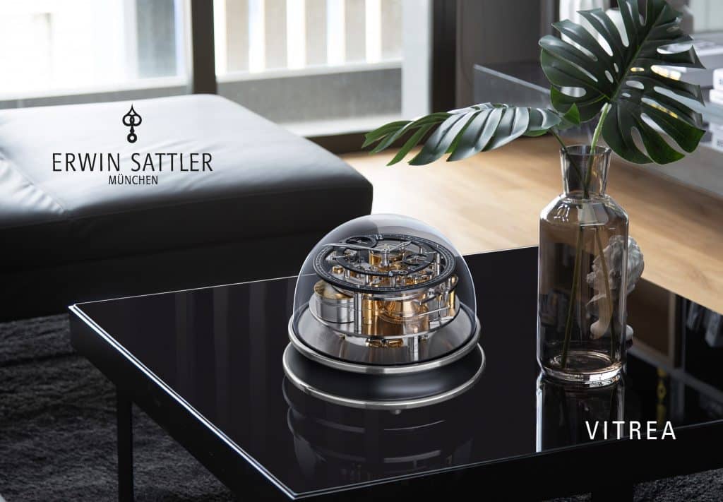 Tischuhr Vitrea von Erwin Sattler mit einem Glasdom und sichtbarem Uhrwerk auf einem schwarzen Glastisch, daneben eine Vase mit großem grünen Blatt. Im Hintergrund ein schwarzes Ledersofa und Regale.