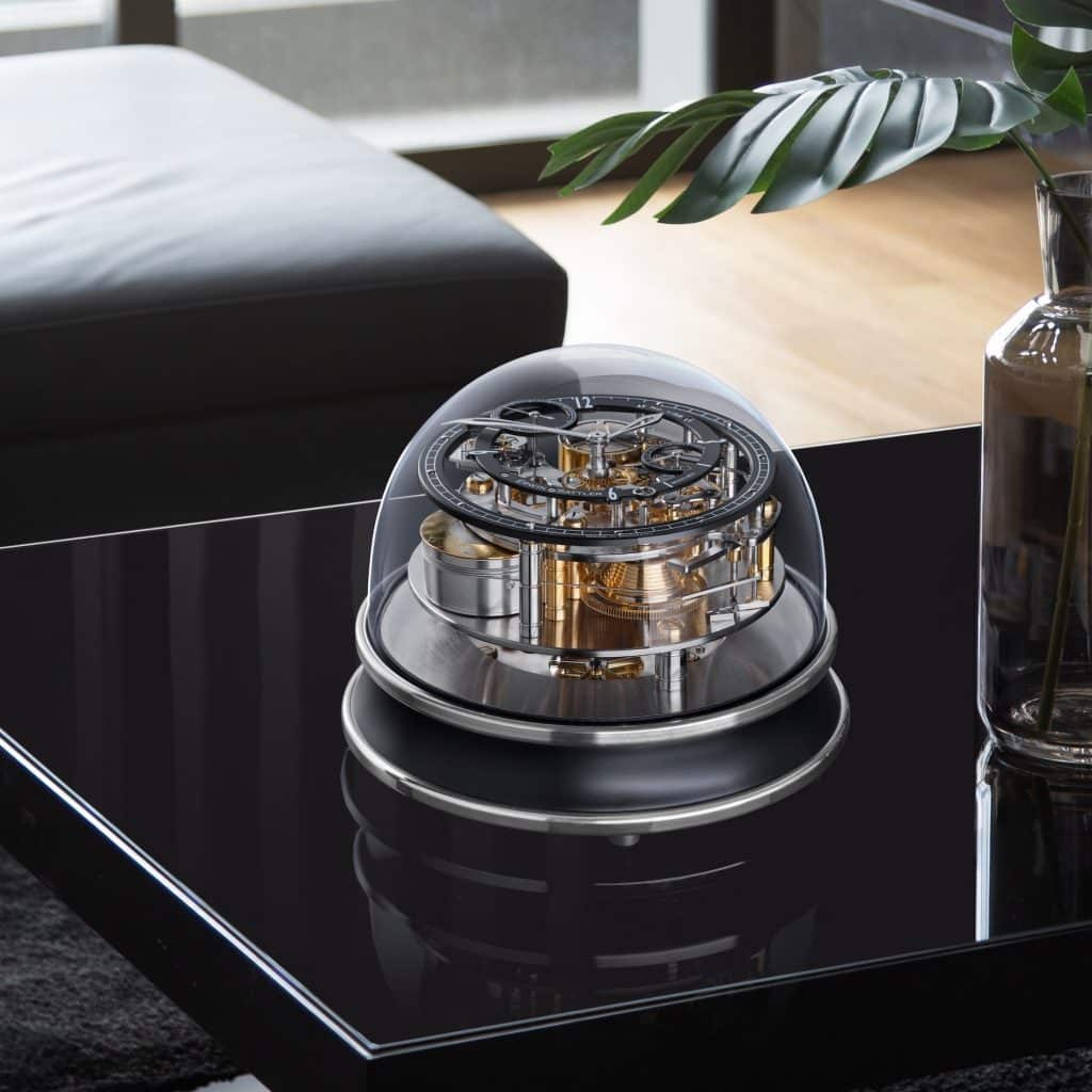 Tischuhr Vitrea von Erwin Sattler auf einem schwarzen Coffeetable daneben steht eine Glasvase und eine kleine Marmosbüste. Eine Ledercouch ist im Hintergrund zu erkennen