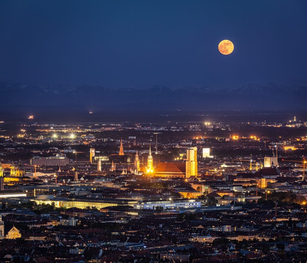 Stadt München bei Nacht mit rotem Blutmond am dunklen Nachthimmel. Stadt mit Frauenkirche und Lichtern