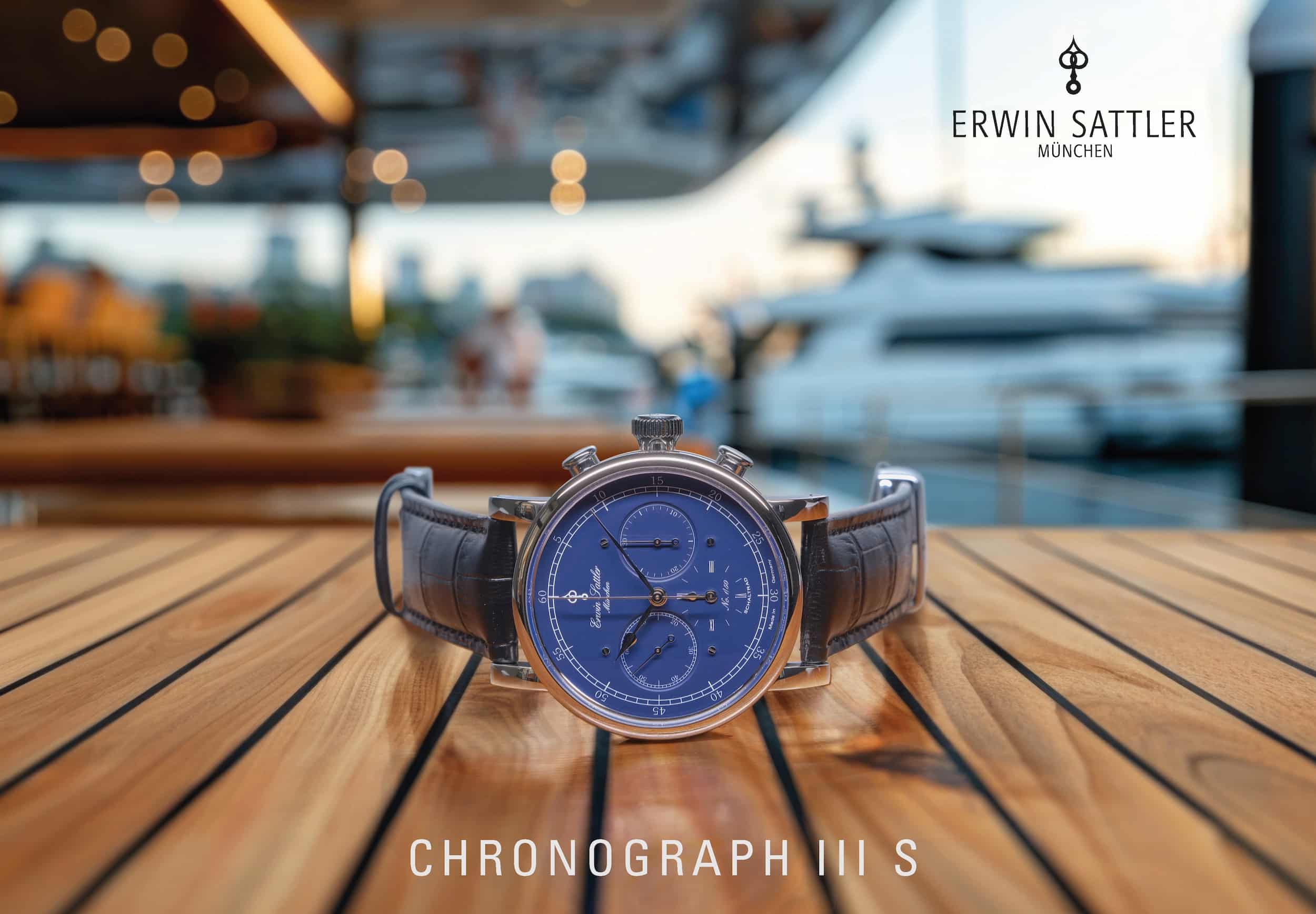 Armbanduhr Chronograph III S von Erwin Sattler mit blauem Zifferblatt und dunkelbraunem Lederarmband auf einem Holztisch, unscharfer maritimer Hintergrund.
